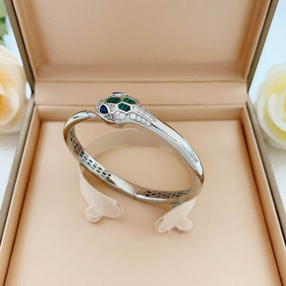 A silver snake ring with green and blue gemstone eyes and clear accent stones, displayed inside a beige jewelry box.