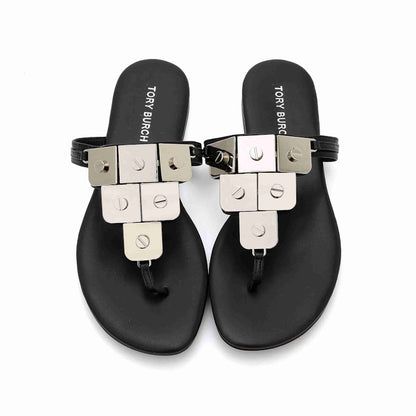 A pair of black flat thong sandals with chunky silver-tone metal block embellishments and black straps, shown from a top-down view on a white background. The insole displays the brand name TORY 