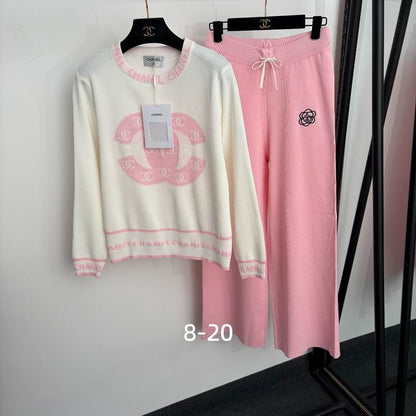 A two-piece loungewear set hanging on black hangers against a white wall. The set includes a cream-colored knit sweater with pink trim and a large pink interlocking CC logo, paired with matching pink ribbed sweatpants featuring a small black CC logo and a white drawstring.
