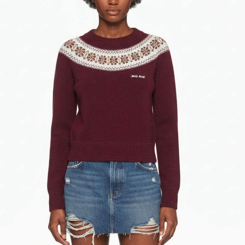 A model wearing a burgundy knit sweater with a white and brown patterned yoke and a small white embroidered logo on the chest, paired with a distressed denim mini skirt, against a plain white background.