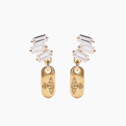 A pair of gold drop earrings featuring a cluster of clear baguette-cut crystals at the top and a polished gold oval pendant below, each engraved with a geometric symbol, set against a plain light gray background.