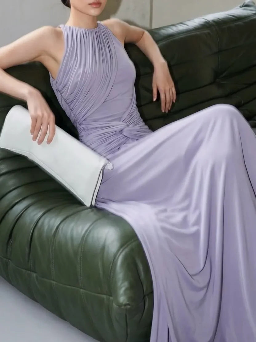 A woman wearing a lavender pleated sleeveless maxi dress sits on a dark green leather sofa, holding a white clutch.