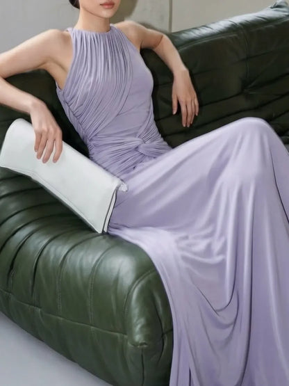 A woman wearing a lavender pleated sleeveless maxi dress sits on a dark green leather sofa, holding a white clutch.