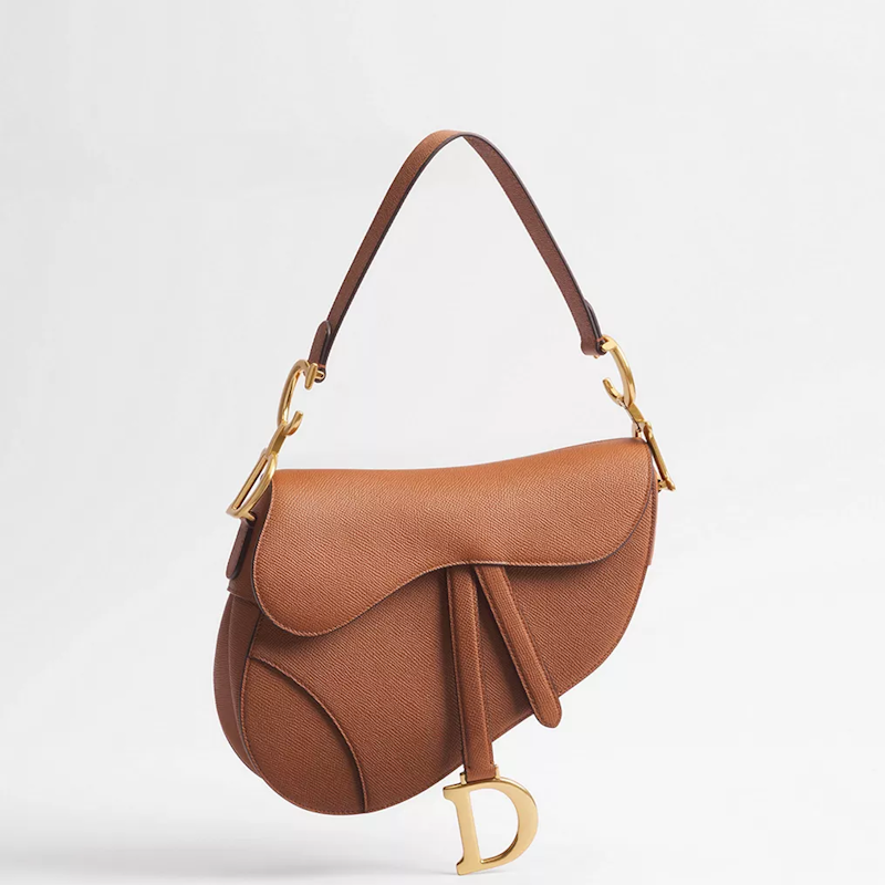 A brown leather saddle bag with gold-tone hardware and a hanging D-shaped charm, photographed against a clean white background.
